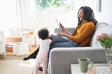 Mother seating on a couch with a table in one hand and playing with her child